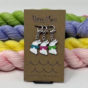 May include: Three white bunny stitch markers with blue, pink, and green bow ties hang from silver clasps on a brown card. The card reads "Three by the Sea designs" and "Stitch Markers". Colorful yarn skeins are in the background.