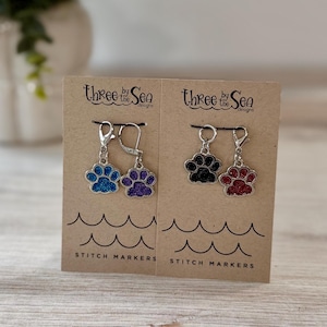 May include: Four metal stitch markers with paw print charms attached to silver clasps, displayed on two brown cards labeled Three by the Sea Designs. The charms feature blue, purple, black, and red glitter finishes. Each card has wave graphics.