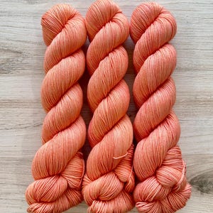 May include: Three skeins of yarn in a vibrant coral-orange hue. Each skein is tightly twisted into a long, cylindrical shape, showcasing a smooth, uniform texture and consistent color throughout the fibers of the twisted yarn strands.