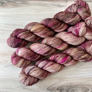 Puede incluir: Madejas de hilo teñido a mano en tonos burdeos, beige y rosa. El hilo está torcido en madejas ordenadas, mostrando las variaciones de color. La textura parece suave e invitante, perfecta para proyectos de tejido o ganchillo.