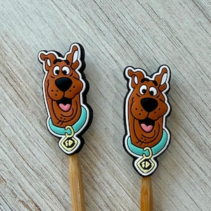 May include: Two brown dog-shaped charms with black outlines, each wearing a light blue collar with a yellow tag featuring the letters SD. The charms are attached to the ends of two light brown wooden sticks, displaying a cartoon character design.