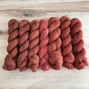 May include: An assortment of yarn skeins in various shades of red and pink. The yarn includes speckled and solid textures, with a fluffy, rose-colored yarn in the center. The skeins are arranged on a light wood surface.