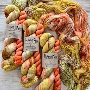 May include: Three skeins of hand-dyed yarn featuring a variegated color palette of yellow, olive green, and coral orange. Each skein has a paper label reading Three by the Sea Designs, Hand-Dyed Yarn, Made in Sunny Florida, with social media handles.