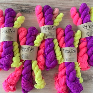 Könnte beinhalten: Drei Knäuel handgefärbtes Garn in leuchtenden Farben Pink, Lila und Gelb. Das Garn ist verdreht und mit "Three by the Sea Hand-Dyed Yarn" beschriftet.