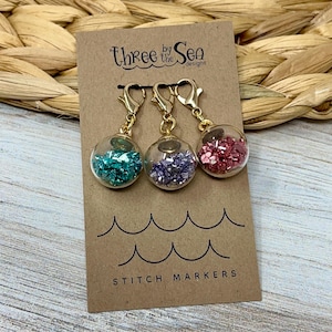 May include: Three by the Sea Designs stitch markers. Set of three gold stitch markers with clear glass beads filled with glitter. The beads are filled with turquoise, purple, and pink glitter.