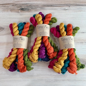 Peut inclure: Trois écheveaux de fil teint à la main, chacun entouré d'une étiquette en papier portant l'inscription Three by the Sea Hand-Dyed Yarn. Le fil présente un mélange de couleurs vives : magenta, jaune moutarde, bleu sarcelle, orange et vert olive.