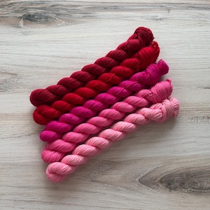 Op de afbeelding: Een assortiment garen in een gradiënt van rode en roze tinten. Het garen is diagonaal gerangschikt en toont een reeks tinten van diep karmozijnrood tot lichtroze. De strengen zijn netjes opgewonden, wat duidt op een knutsel- of breiproject.
