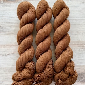 May include: Three skeins of medium brown yarn, each twisted into a long, uniform coil. The yarn features a smooth, consistent texture and a rich, warm earth-toned color throughout, with each skein displaying a tightly wound, multi-ply structure.
