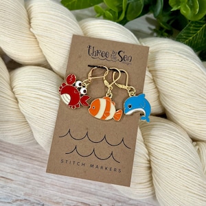 May include: Three by the Sea Designs stitch markers featuring a red crab, an orange and white clownfish, and a blue dolphin. The markers are on a brown card with the text "Three by the Sea Designs" and "Stitch Markers".