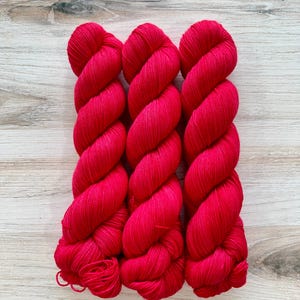 May include: Three skeins of vibrant red yarn are arranged vertically on a light wood surface. The yarn is tightly twisted, showcasing its texture and color. Ideal for knitting or crochet projects.