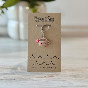 Può includere: Un marcapunti in metallo color argento con chiusura a leva fissato su un cartoncino color marrone chiaro. Il ciondolo è una piccola tazza da caffè beige sorridente con un minuscolo cuore rosa sul lato. Il cartoncino riporta il testo Three by the Sea Designs e STITCH MARKERS.
