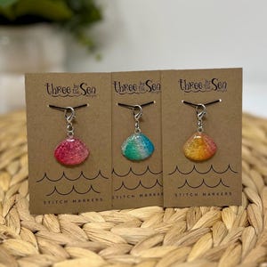 May include: Three stitch markers in the shape of seashells, each attached to a small card. The shells are in shades of pink, blue-green, and yellow-orange. The cards have the text "Three by the Sea" and "STITCH MARKERS".
