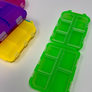 Puede incluir: Una colecci&oacute;n de pastilleros de pl&aacute;stico de colores verde, amarillo, morado y rosa. El organizador verde abierto muestra m&uacute;ltiples compartimentos. Los organizadores tienen forma rectangular con tapa abatible y cierres seguros.
