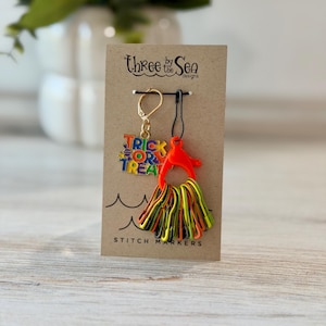 May include: A set of stitch markers on a brown display card labeled Three by the Sea Designs. The set includes a gold-tone charm with multi-colored text reading Trick or Treat and a collection of orange, yellow, and black safety pin markers.