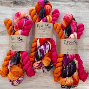 May include: Three skeins of hand-dyed yarn featuring a vibrant variegated color palette of hot pink, bright orange, white, and deep charcoal gray. Each skein is wrapped in a tan paper label printed with the text Three by the Sea Designs.