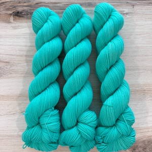 May include: Three skeins of turquoise yarn are arranged vertically against a light wood grain surface. The yarn is tightly wound, showcasing a vibrant, solid color. This image is ideal for knitters and crocheters.