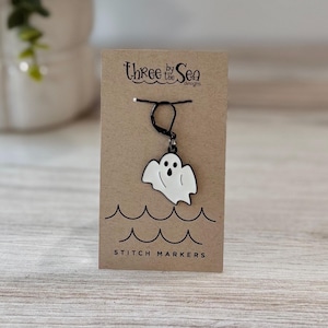 May include: A white ghost-shaped enamel stitch marker with black outlines and facial features, attached to a black metal lever-back clasp. The marker is displayed on a rectangular kraft paper card printed with the text Three by the Sea designs and STITCH MARKERS above and below a graphic of black wavy lines.
