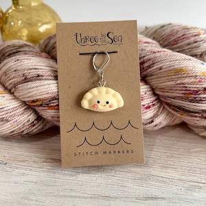 Puede incluir: Un marcador de punto con forma de dumpling sonriente, sujeto a un cierre plateado, cuelga de una tarjeta marrón. La tarjeta tiene el texto "Three by the Sea designs" y "STITCH MARKERS". El fondo incluye un ovillo de lana.
