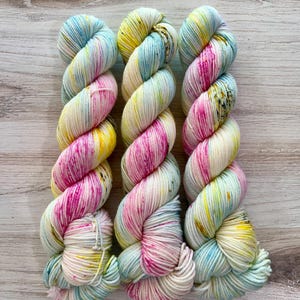 Puede incluir: Tres madejas de hilo multicolor, dispuestas verticalmente. El hilo presenta una mezcla de blanco, rosa, amarillo y azul, con un aspecto moteado. El hilo está torcido en un patrón similar a una cuerda, ideal para proyectos de tejido o ganchillo.