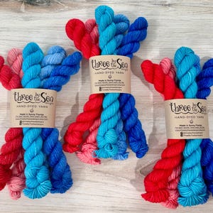 May include: Three bundles of hand-dyed yarn, each featuring a gradient of red, pink, light blue, and dark blue strands. Each bundle is secured with a tan paper label printed with the text Three by the Sea Hand-Dyed Yarn, Made in Sunny Florida.