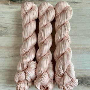 May include: Three skeins of blush pink yarn are displayed against a light wood grain surface. The yarn is tightly twisted into a rope-like form, with a soft, matte finish. The color is a delicate, muted pink, suitable for knitting or crochet projects.