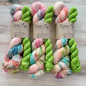 Peut inclure: Trois écheveaux de fil teints à la main aux couleurs rose, vert et bleu. Chaque écheveau est étiqueté avec "Three by the Sea" et "Hand-Dyed Yarn". Le fil est disposé sur une surface en bois clair.