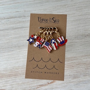 Peut inclure: Un ensemble de cinq marqueurs de mailles en métal sur le thème patriotique fixés sur une carte brune étiquetée Three by the Sea Designs. Les breloques présentent des motifs en émail rouge, blanc et bleu incluant une étoile, USA, un drapeau, une tong et un haut-de-forme.