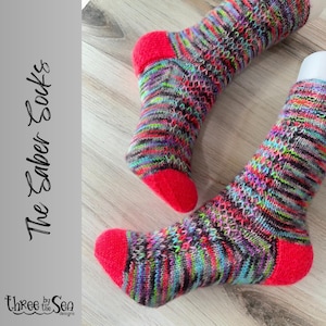 May include: A pair of colorful knit socks with a red heel and toe. The socks have a multi-colored pattern with shades of pink, blue, green, and black. The socks are called "The Ember Socks" by "Three by the Sea Designs".