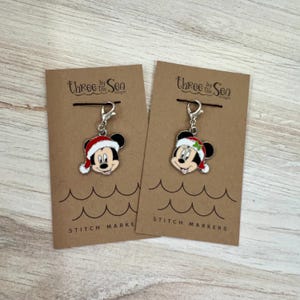 May include: Two Mickey Mouse stitch markers with a Christmas theme. Each marker features a Mickey Mouse head wearing a Santa hat. The markers are attached to a silver clasp and displayed on a brown card with the text "Three by the Sea" and "Stitch Markers".