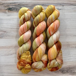 May include: Three skeins of hand-dyed yarn arranged vertically. The yarn features a variegated color palette of warm yellow, soft orange, muted olive green, and cream white tones, twisted into a tight, uniform spiral structure for display.