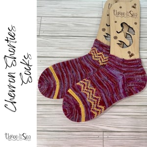 May include: A pair of chevron shortie socks in shades of burgundy, purple, and yellow. The socks feature a zig-zag pattern and are displayed on wooden sock forms. The text "Chevron Shorties Socks" is on the left.