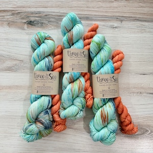 May include: Three skeins of hand-dyed yarn in shades of teal, orange, and brown. The yarn is labeled "Three by the Sea" and includes the text "HAND-DYED YARN". The yarn is arranged on a light wood surface.