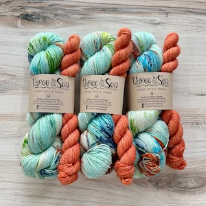May include: Six skeins of hand-dyed yarn in shades of teal, orange, and blue. The yarn is labeled "Three by the Sea Designs" and includes the text "Made in Sunny Florida". The yarn is arranged on a light wood surface.