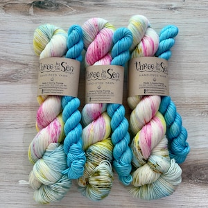 May include: Three skeins of hand-dyed yarn in shades of white, pink, yellow, and turquoise. Each skein is labeled with "Three by the Sea" and "HAND-DYED YARN". The yarn is arranged on a light wood surface.