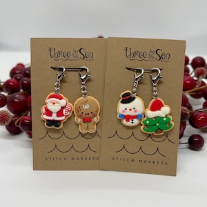 May include: Two sets of Christmas-themed stitch markers. One set features Santa Claus and a gingerbread man, the other a snowman and a Christmas tree. Each marker is attached to a silver-colored clasp and displayed on a brown card with the text "Three by the Sea" and "Stitch Markers".