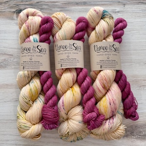 May include: Three skeins of hand-dyed yarn with a cream base and speckles of yellow, teal, and pink, each paired with a solid magenta mini-skein. Each bundle features a paper label reading Three by the Sea Designs Hand-Dyed Yarn Made in Sunny Florida.