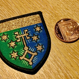 May include: A green and blue embroidered patch with gold glitter accents. The patch features a stylised cross with stars around it. The patch is shaped like a shield.