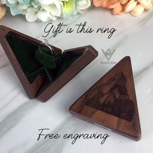 May include: A wooden triangular ring box with a hinged lid, open to reveal a ring on a green velvet insert. The lid is engraved with a mountain scene. Text on the image reads "Gift is this ring" and "Free engraving."