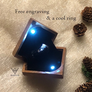 May include: A wooden ring box with a black velvet interior and a silver ring with a clear stone. The box is open and lit from inside, with the text "Free engraving & a cool ring" above the box.