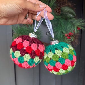 May include: Two handmade crochet Christmas baubles. One is burgundy, red, and pink, the other is green, red, and pink. Each has a silver cap and a white ribbon for hanging. The baubles are held in front of a Christmas wreath.