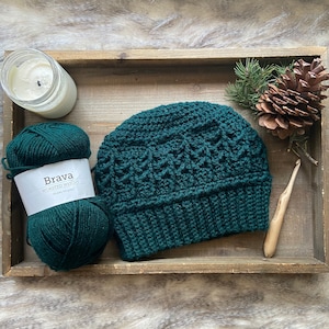 May include: A dark green crocheted beanie hat with a textured pattern. The hat is sitting on a wooden surface with a skein of dark green yarn and a crochet hook.