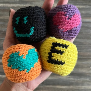 Puede incluir: Cuatro bolas de crochet con diferentes diseños. Una bola es negra con una cara sonriente blanca, una es morada con un corazón rosa, una es naranja con un corazón turquesa y una es amarilla con una letra E negra.