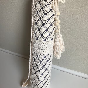 May include: A white crocheted yoga mat bag with a drawstring closure and a tassel. The bag has a white and cream color scheme and is made with a delicate crochet pattern.