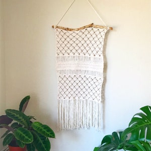 May include: White macrame wall hanging with a geometric pattern and fringe. The hanging is attached to a natural wood branch.