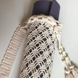 May include: A white crocheted yoga mat bag with a long strap and tassels. The bag is holding a rolled up purple yoga mat.