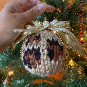 May include: A brown, black, and beige crocheted Christmas ornament with a gold and white ribbon. The ornament is shaped like a ball and has a leopard print pattern.