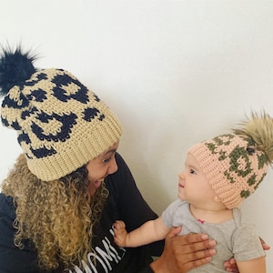 May include: A woman and a baby wearing matching crocheted hats with a leopard print design. The woman's hat is beige with black spots, and the baby's hat is pink with green spots. Both hats have black pom-poms.