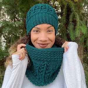 Puede incluir: Un gorro y un cuello a juego de punto de ganchillo de color verde oscuro. El gorro tiene un diseño texturizado y el cuello es un punto sencillo.