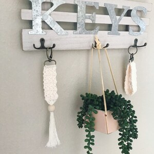 May include: A white wooden sign with the word "KEYS" in metal letters. There are three hooks on the sign, with a white crocheted key chain with a wooden bead and tassel hanging from one hook, a hanging planter with a succulent plant hanging from another hook, and a white crocheted key chain hanging from the third hook.