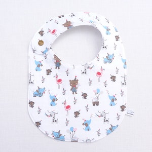 Könnte beinhalten: Ein weißes Lätzchen für Babys mit einem wiederkehrenden Muster aus bunten Zeichentricktieren, darunter braune Bären, blaue Elefanten, rosa Flamingos und Affen mit Partyhüten. Das Lätzchen hat einen braunen Druckknopf am Hals und ein Etikett.